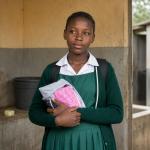 A school girl, holding a menstrual hygiene kit near a modest latrine facility.