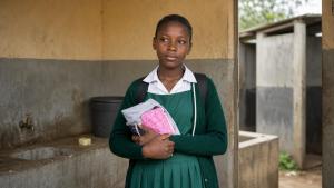 A school girl, holding a menstrual hygiene kit near a modest latrine facility.